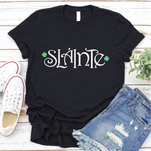 Puede incluir: Camiseta negra con la frase irlandesa "Sl&aacute;inte" en letras blancas y tr&eacute;boles verdes a cada lado.
