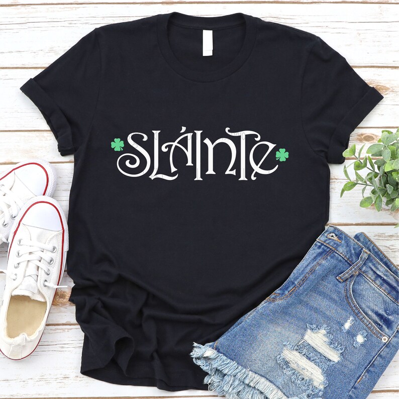 Puede incluir: Camiseta negra con la frase irlandesa "Sl&aacute;inte" en letras blancas y tr&eacute;boles verdes a cada lado.