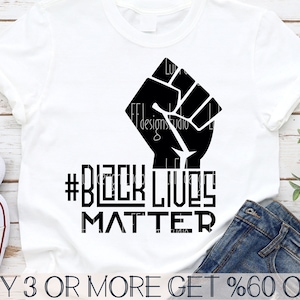 May include: A white t-shirt with a black raised fist and the text "#BLACK LIVES MATTER" printed on it. The t-shirt is on a wooden surface with a pair of jeans and a plant in the background.