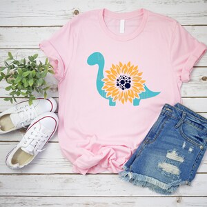 May include: A pink t-shirt with a teal dinosaur silhouette with a yellow sunflower design in the center. The sunflower has a dark blue center with white dots.