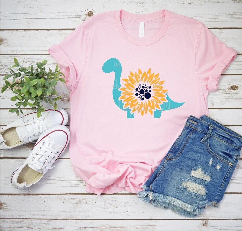 May include: A pink t-shirt with a teal dinosaur silhouette with a yellow sunflower design in the center. The sunflower has a dark blue center with white dots.