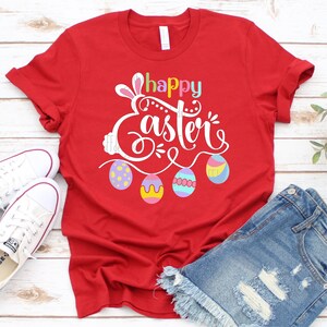 May include: Red t-shirt with a white graphic that says "happy easter" with a bunny ear design and colorful Easter eggs.