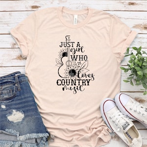 Puede incluir: Camiseta rosa claro con el texto negro "Just a girl who loves country music". El dise&ntilde;o presenta una silueta de guitarra con girasoles y hojas. La camiseta se muestra con pantalones cortos de mezclilla y zapatillas blancas.