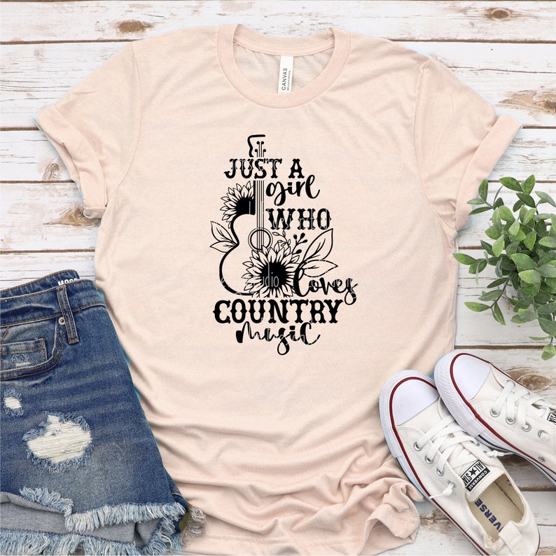 Puede incluir: Camiseta rosa claro con el texto negro "Just a girl who loves country music". El dise&ntilde;o presenta una silueta de guitarra con girasoles y hojas. La camiseta se muestra con pantalones cortos de mezclilla y zapatillas blancas.