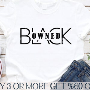 May include: White t-shirt with the text "BLACK OWNED" in black lettering. The text is in a bold, blocky font. The shirt is laying on a wooden surface with a pair of jeans and a pair of sneakers.