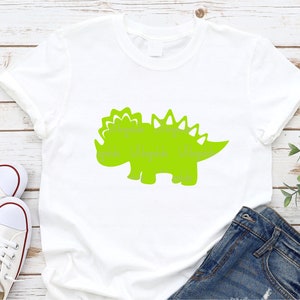以下が含まれることがあります： 緑色の恐竜のシルエットのデザインが施された白いTシャツ。恐竜は背中にトゲがあり、頭にはフリルがあります。