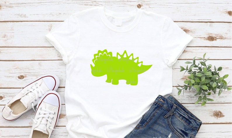 以下が含まれることがあります： 緑色の恐竜のシルエットのデザインが施された白いTシャツ。恐竜は背中にトゲがあり、頭にはフリルがあります。