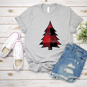 May include: A gray t-shirt with a red and black buffalo plaid Christmas tree design. The shirt is laid flat on a wooden surface with a pair of white sneakers and denim shorts.