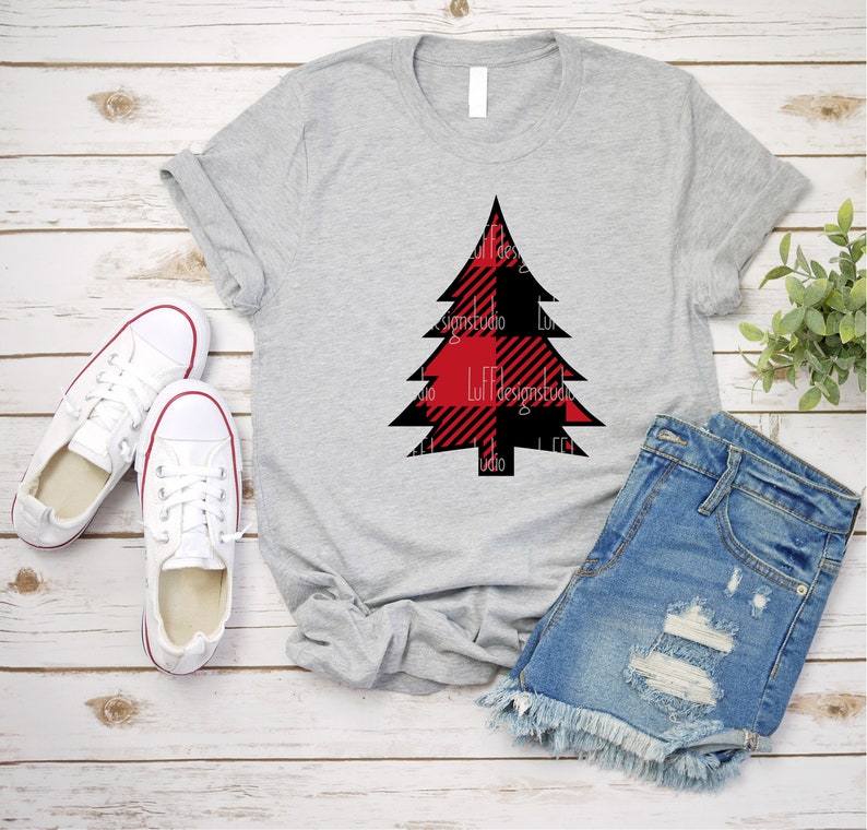 May include: A gray t-shirt with a red and black buffalo plaid Christmas tree design. The shirt is laid flat on a wooden surface with a pair of white sneakers and denim shorts.