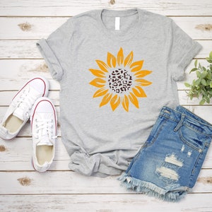 May include: A gray t-shirt with a yellow sunflower graphic. The center of the sunflower is a leopard print pattern. The t-shirt is laying on a white wood background with a pair of white sneakers and denim shorts.