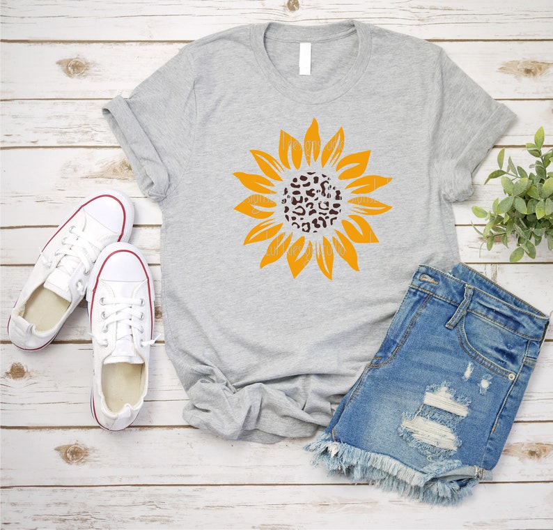 May include: A gray t-shirt with a yellow sunflower graphic. The center of the sunflower is a leopard print pattern. The t-shirt is laying on a white wood background with a pair of white sneakers and denim shorts.