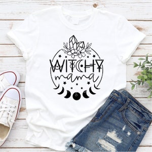 May include: White t-shirt with a black graphic design that says "Witchy Mama" in a circle with stars, moons, and crystals.