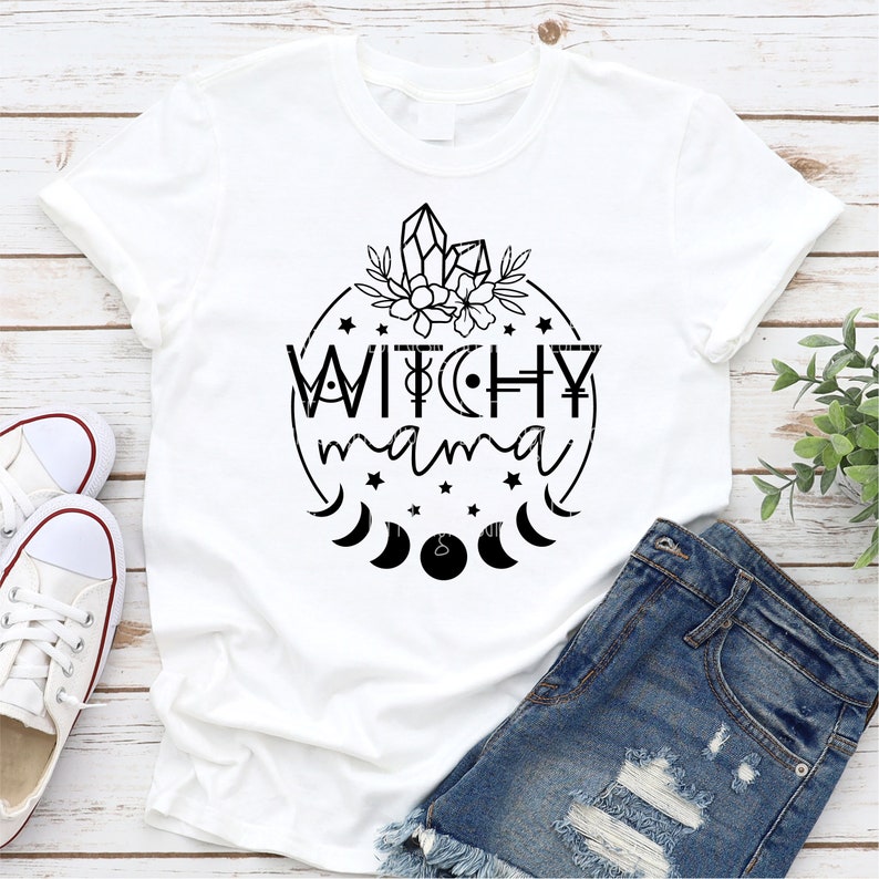 May include: White t-shirt with a black graphic design that says "Witchy Mama" in a circle with stars, moons, and crystals.