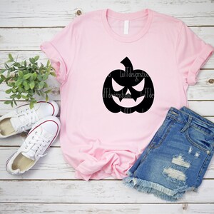Op de afbeelding: Een roze T-shirt met een zwart silhouet van een jack-o'-lantern met een dreigende grijns. Het T-shirt ligt op een wit houten oppervlak met een paar witte sneakers en een blauwe spijkerbroek.