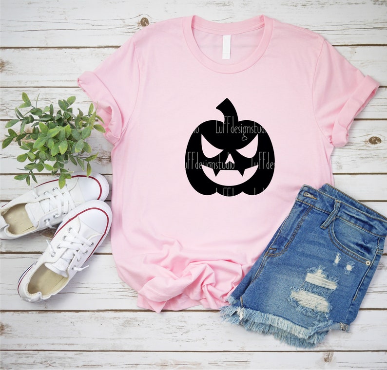 Op de afbeelding: Een roze T-shirt met een zwart silhouet van een jack-o'-lantern met een dreigende grijns. Het T-shirt ligt op een wit houten oppervlak met een paar witte sneakers en een blauwe spijkerbroek.
