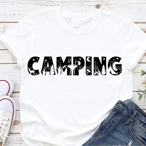 Op de afbeelding: Wit T-shirt met zwarte tekst die "CAMPING" zegt met camping-silhouetten in de letters.