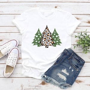 K&ouml;nnte beinhalten: Wei&szlig;es T-Shirt mit einer Grafik von drei Weihnachtsb&auml;umen. Der mittlere Baum hat ein Leopardenmuster, flankiert von zwei gr&uuml;nen B&auml;umen. Das Shirt wird mit Jeansshorts und wei&szlig;en Turnschuhen kombiniert, was einen l&auml;ssigen, festlichen Look ergibt.