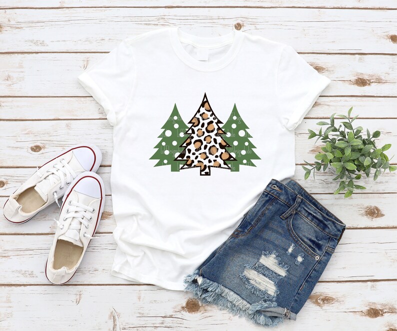 K&ouml;nnte beinhalten: Wei&szlig;es T-Shirt mit einer Grafik von drei Weihnachtsb&auml;umen. Der mittlere Baum hat ein Leopardenmuster, flankiert von zwei gr&uuml;nen B&auml;umen. Das Shirt wird mit Jeansshorts und wei&szlig;en Turnschuhen kombiniert, was einen l&auml;ssigen, festlichen Look ergibt.