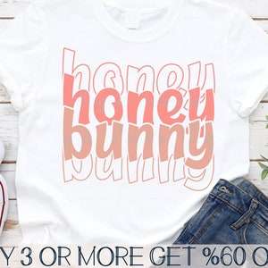 May include: A white t-shirt with the text "honey bunny" in pink and white lettering. The text is in a retro style font. The shirt is on a wooden surface with a pair of jeans and a pair of white sneakers.