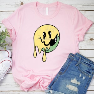 May include: A pink t-shirt featuring a yellow and green dripping smiley face graphic. The graphic has a black outline and the smiley face has black eyes and a black mouth.