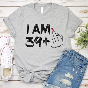 May include: Gray t-shirt with the text "I AM 39+" and a black and white illustration of a hand giving the middle finger. The hand has red nail polish.