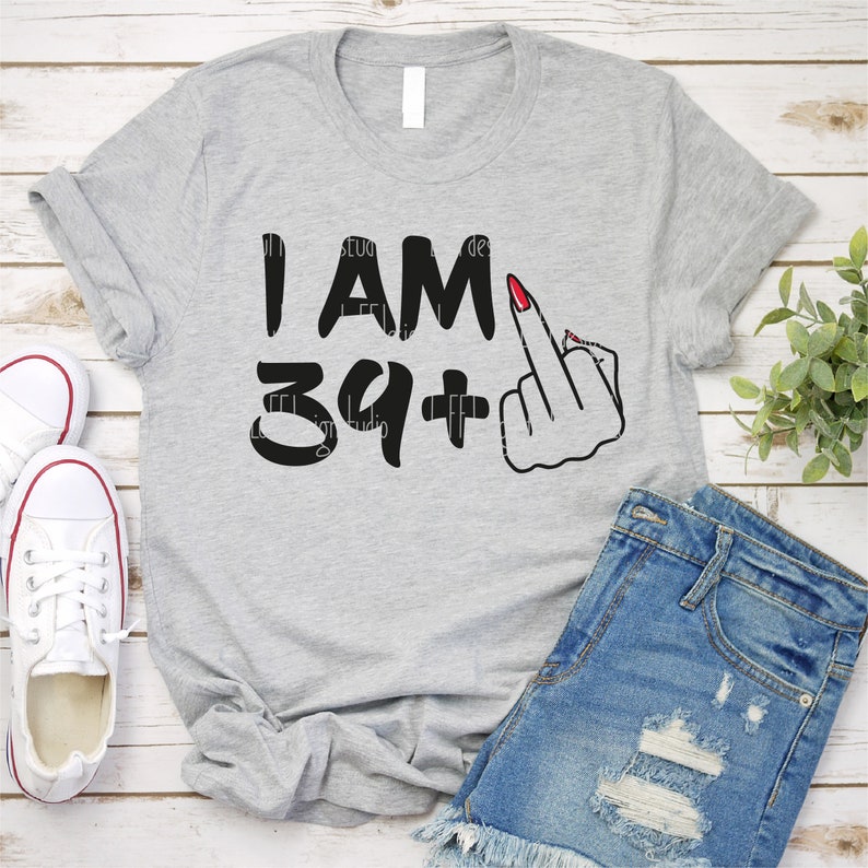 May include: Gray t-shirt with the text "I AM 39+" and a black and white illustration of a hand giving the middle finger. The hand has red nail polish.