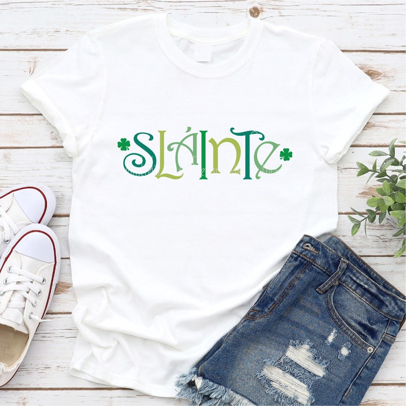 Puede incluir: Camiseta blanca con la palabra "Sl&aacute;inte" en letras verdes, amarillas y azules con acentos de tr&eacute;bol.