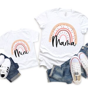 K&ouml;nnte beinhalten: Ein Set aus zwei wei&szlig;en T-Shirts mit einem Regenbogen-Design und den Worten "Mama" und "Mini" darauf gedruckt. Die Shirts liegen auf einem wei&szlig;en Hintergrund mit blauen Jeans und wei&szlig;en Turnschuhen.
