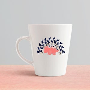 May include: White ceramic mug with a pink dinosaur silhouette under a navy blue leafy archway design.