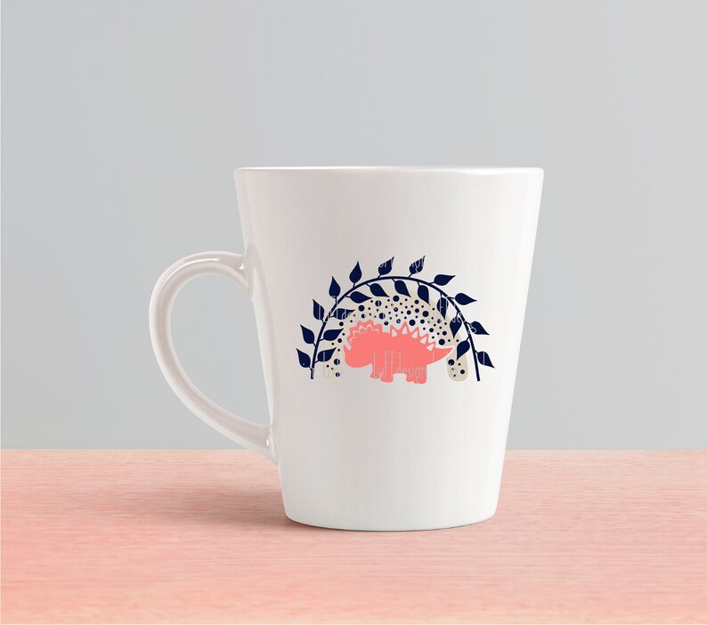 May include: White ceramic mug with a pink dinosaur silhouette under a navy blue leafy archway design.