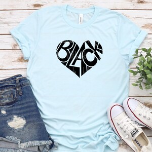 Puede incluir: Camiseta azul claro con un dise&ntilde;o de coraz&oacute;n negro. La palabra "BLACK" est&aacute; formada dentro del coraz&oacute;n. La camiseta se combina con pantalones cortos de mezclilla y zapatillas de lona blancas.