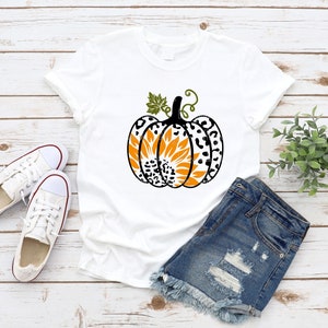 K&ouml;nnte beinhalten: Wei&szlig;es T-Shirt mit einem schwarzen und wei&szlig;en Leopardenmuster-K&uuml;rbis mit einer Sonnenblume im Inneren. Der K&uuml;rbis hat einen gr&uuml;nen Stiel und Bl&auml;tter.