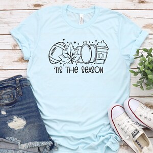 May include: A light blue t-shirt with a black graphic design of a football, a leaf, a pumpkin, and a coffee cup. The text "'Tis the season" is printed below the graphic.