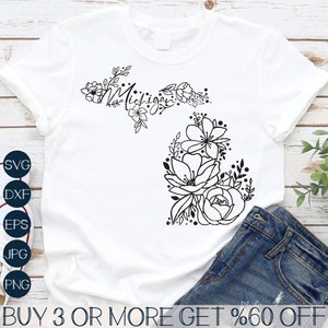 Könnte beinhalten: Ein weißes T-Shirt mit einer schwarzen Umrandung des Bundesstaates Michigan mit floralen Designs und dem Wort "Michigan" innerhalb der Umrandung. Das T-Shirt liegt auf einer Holzoberfläche mit einer blauen Jeans.