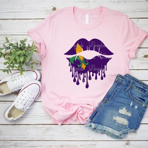 May include: A pink t-shirt with a purple and gold dripping lips design featuring a fleur-de-lis. The shirt is laid out on a wooden surface with a pair of white sneakers and blue denim shorts.