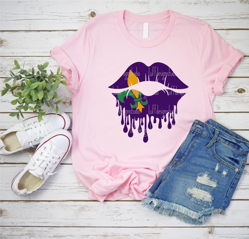 May include: A pink t-shirt with a purple and gold dripping lips design featuring a fleur-de-lis. The shirt is laid out on a wooden surface with a pair of white sneakers and blue denim shorts.