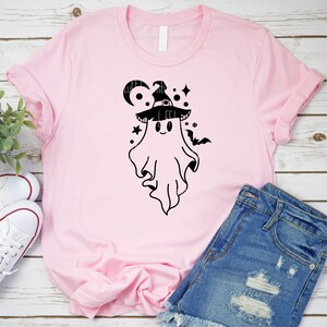 May include: A pink t-shirt with a black graphic of a ghost wearing a witch hat. The ghost has a cute, cartoon-like face and is surrounded by stars and a crescent moon.