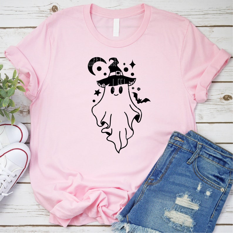 May include: A pink t-shirt with a black graphic of a ghost wearing a witch hat. The ghost has a cute, cartoon-like face and is surrounded by stars and a crescent moon.