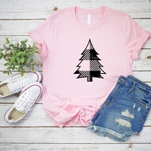 May include: A pink t-shirt with a black and white plaid Christmas tree design. The shirt is laying on a white wooden surface with a pair of white sneakers and a pair of blue denim shorts.
