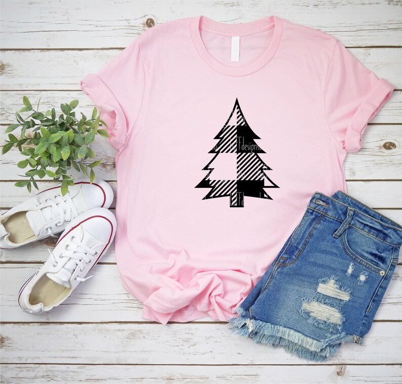 May include: A pink t-shirt with a black and white plaid Christmas tree design. The shirt is laying on a white wooden surface with a pair of white sneakers and a pair of blue denim shorts.