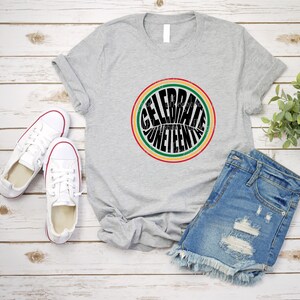 K&ouml;nnte beinhalten: Ein graues T-Shirt mit einem schwarzen Kreis-Motiv, auf dem in einer Retro-Schriftart "Celebrate Juneteenth" steht. Der Kreis ist mit roten, gelben und gr&uuml;nen Streifen umrandet.