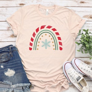 May include: A light pink t-shirt with a red and green candy cane rainbow design and a snowflake in the center. The rainbow is outlined in gold with gold polka dots.