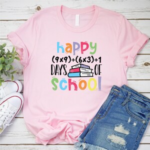 May include: Pink t-shirt with the text "happy (9x9)+(6x3)+1 days of school!" in colorful letters. A stack of books is in the middle of the text.