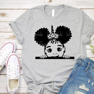 May include: A grey t-shirt featuring a black and white illustration of a girl with afro puffs, a unicorn horn, and a flower headband peeking over the top of a white rectangle.