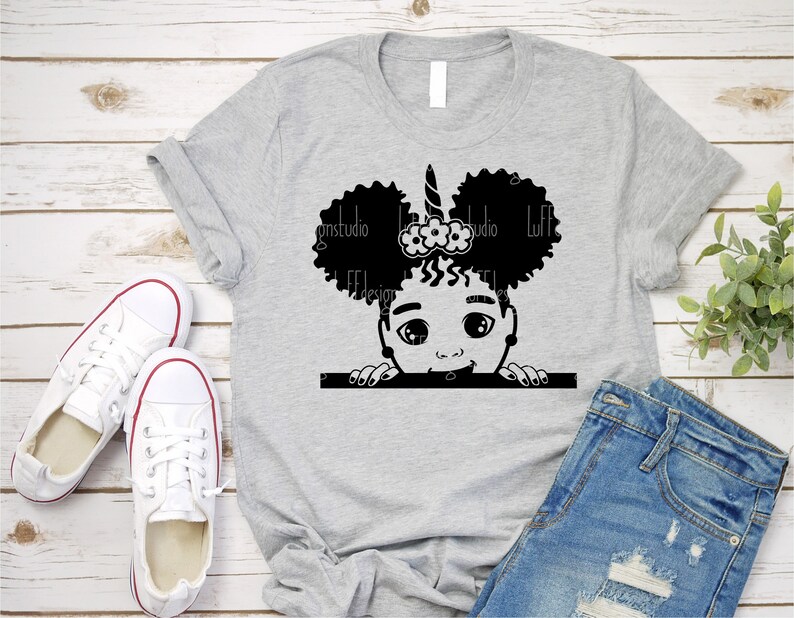 May include: A grey t-shirt featuring a black and white illustration of a girl with afro puffs, a unicorn horn, and a flower headband peeking over the top of a white rectangle.