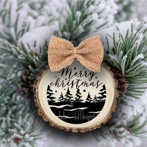 May include: A round wooden ornament with a rustic bark edge. The ornament features a black silhouette of a forest scene with the text "Merry Christmas" and stars.