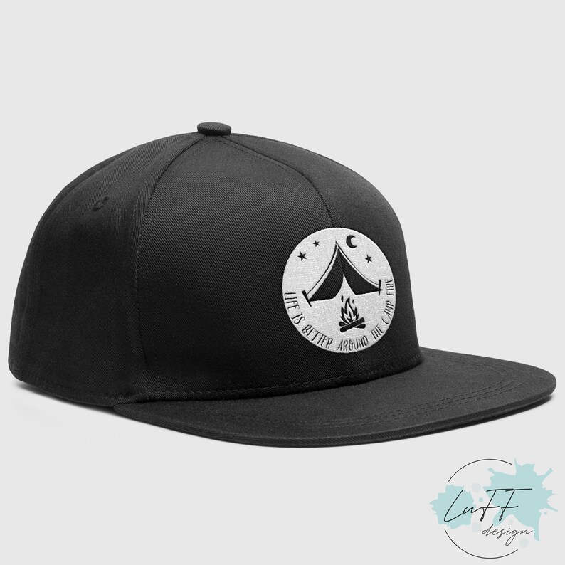 Op de afbeelding: Zwarte en witte geborduurde flat brim baseballcap met een wit cirkelontwerp met een tent, kampvuur, sterren en de tekst "Life is better around the campfire".