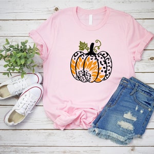 K&ouml;nnte beinhalten: Ein pinkfarbenes T-Shirt mit einem schwarzen und orangefarbenen K&uuml;rbisdesign. Der K&uuml;rbis hat ein Sonnenblumenmuster und Leopardenmuster-Akzente.