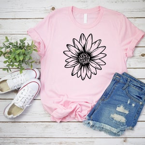 May include: A pink t-shirt with a black daisy graphic design. The t-shirt is laid out on a white wooden surface with a pair of white sneakers and denim shorts.