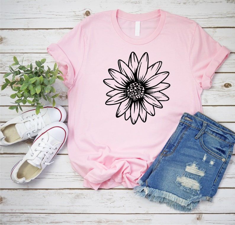 May include: A pink t-shirt with a black daisy graphic design. The t-shirt is laid out on a white wooden surface with a pair of white sneakers and denim shorts.